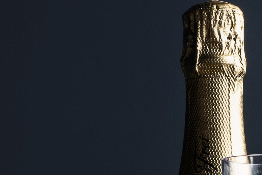 Close-up of the neck of a champagne bottle with a textured gold foil capsule, set against a very dark navy background with a glass partially visible in the lower right corner.