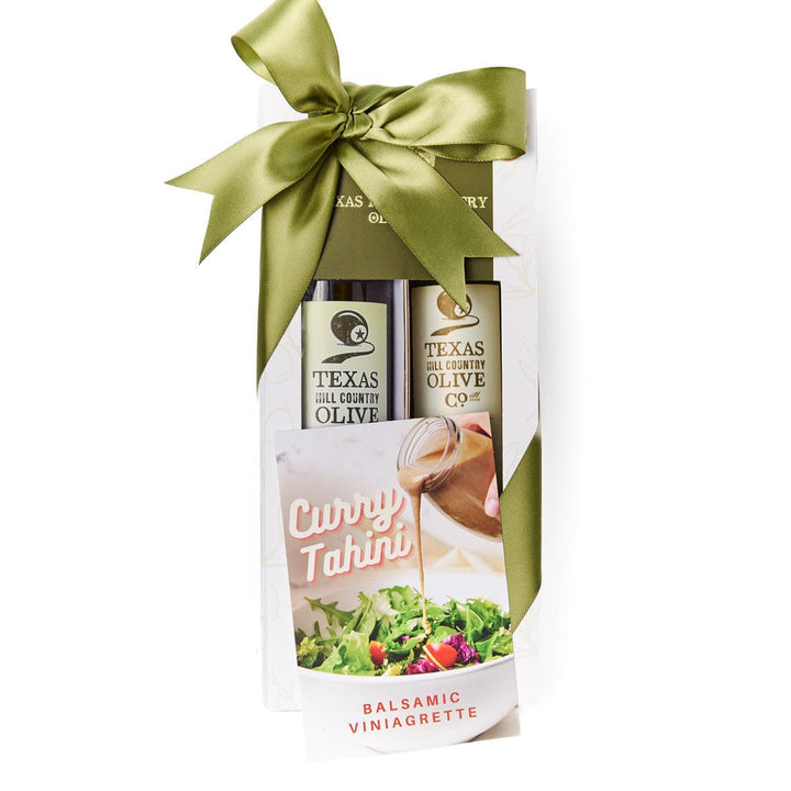A Texas Olive Company gift set in a clear window box package tied with a large olive green satin bow. The set includes a bottle of The Spaniard Olive Oil and White balsamic with Curry Tahini recipe card
