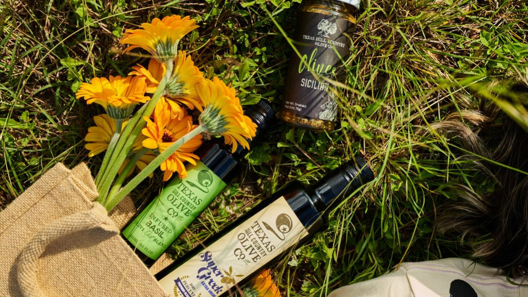 A styled flat-lay of Texas Hill Country Olive Co. products on a grassy lawn with yellow flowers.