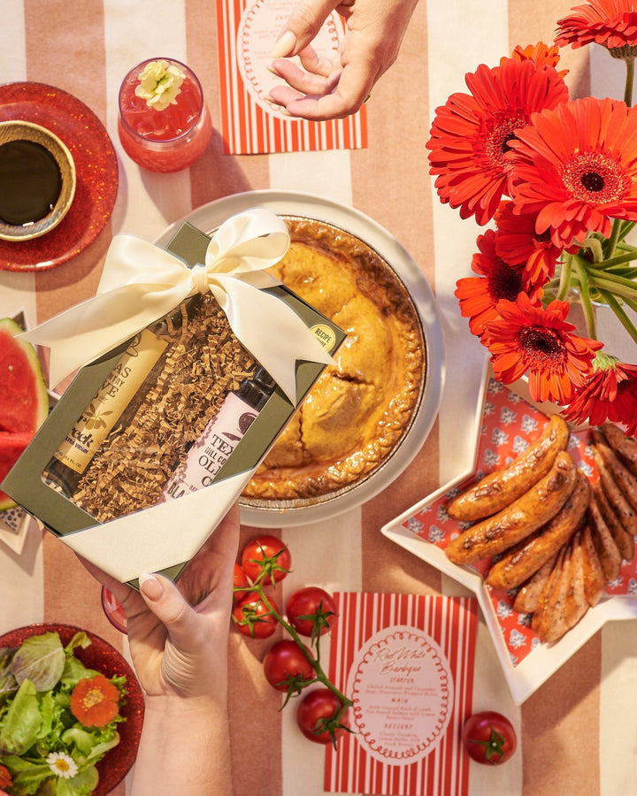 Table setting with a pie, flowers, and various food items on a checkered tablecloth. Passing green gift box with white ribbon across the table.