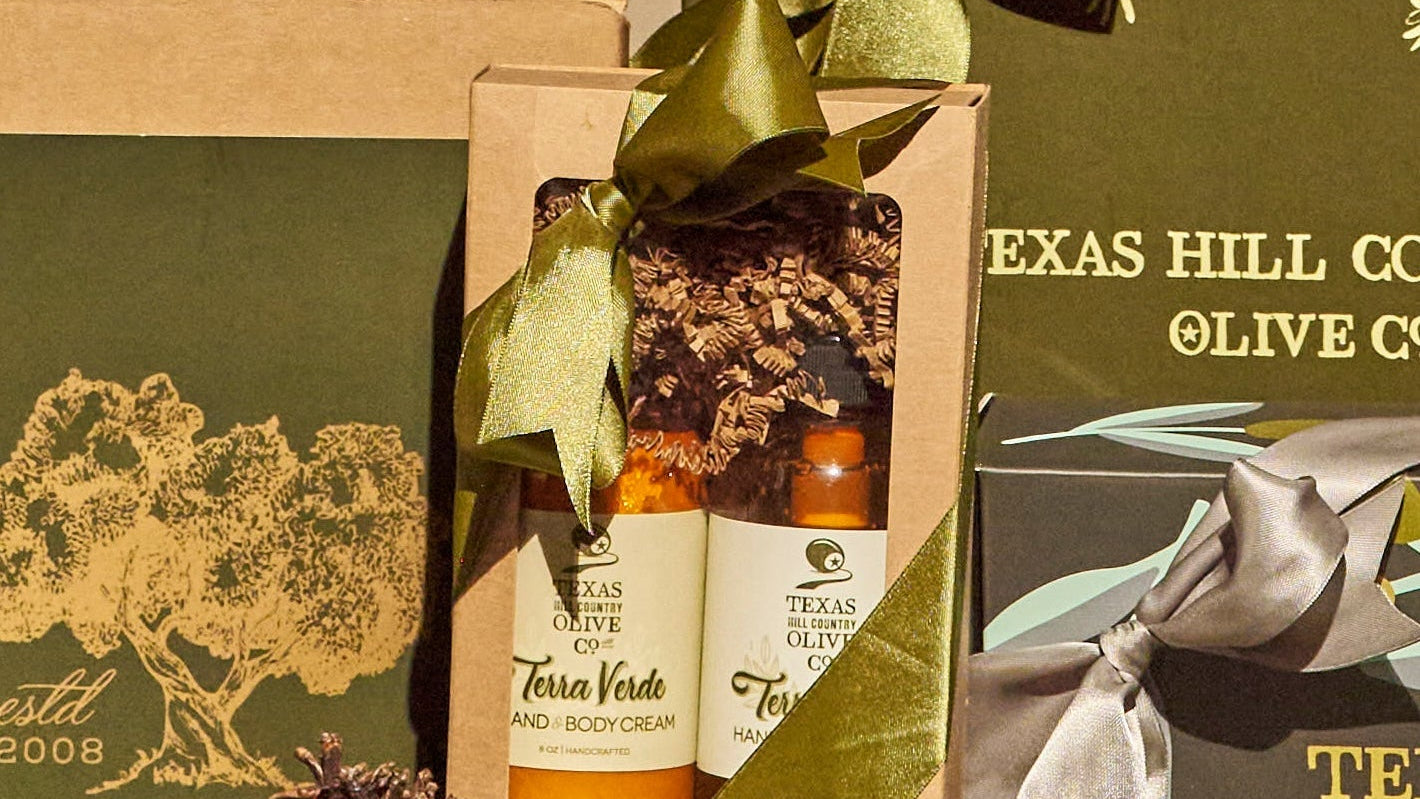 A gift set from Texas Hill Country Olive Co. featuring two small bottles of body cream in a kraft paper box with a green ribbon, surrounded by olive-themed packaging and a pinecone.
