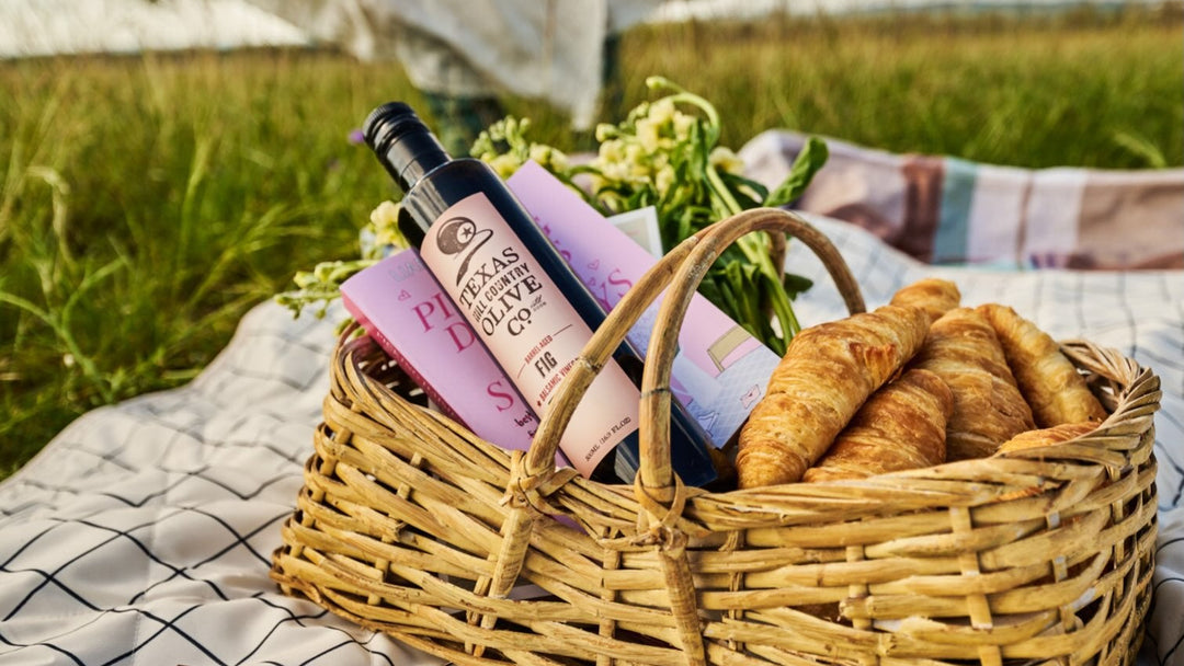 A wicker picnic basket on a white picnic blanket in a grassy field, filled with croissants, flowers, and a bottle of olive oil next to a checkers board.