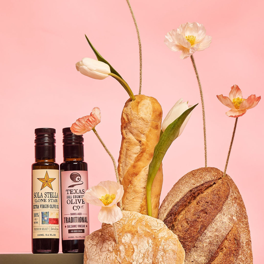 100 ml Sola Stella olive oil and Traditional balsamic bottles displayed with artisan bread and flowers against pink.
