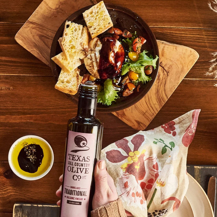 Traditional Aged Balsamic Vinegar beside bread, salad and cheese drizzled with balsamic on a wooden table.
